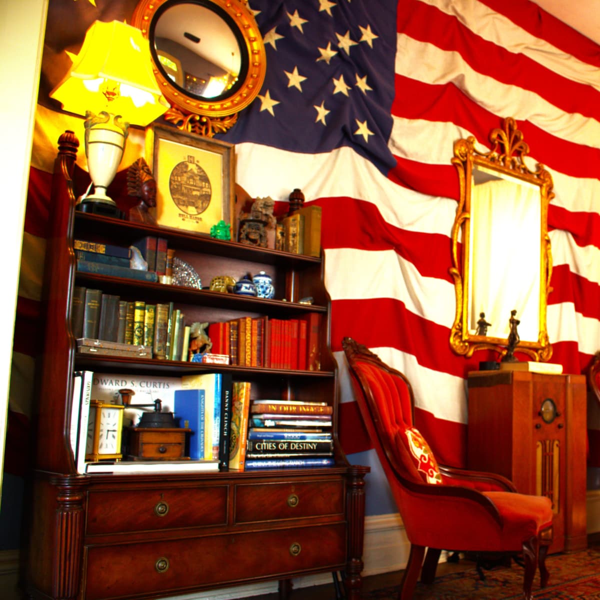 A room decorated with an American flag wall, featuring vintage furniture including a mirror, bookshelves, and two red chairs.