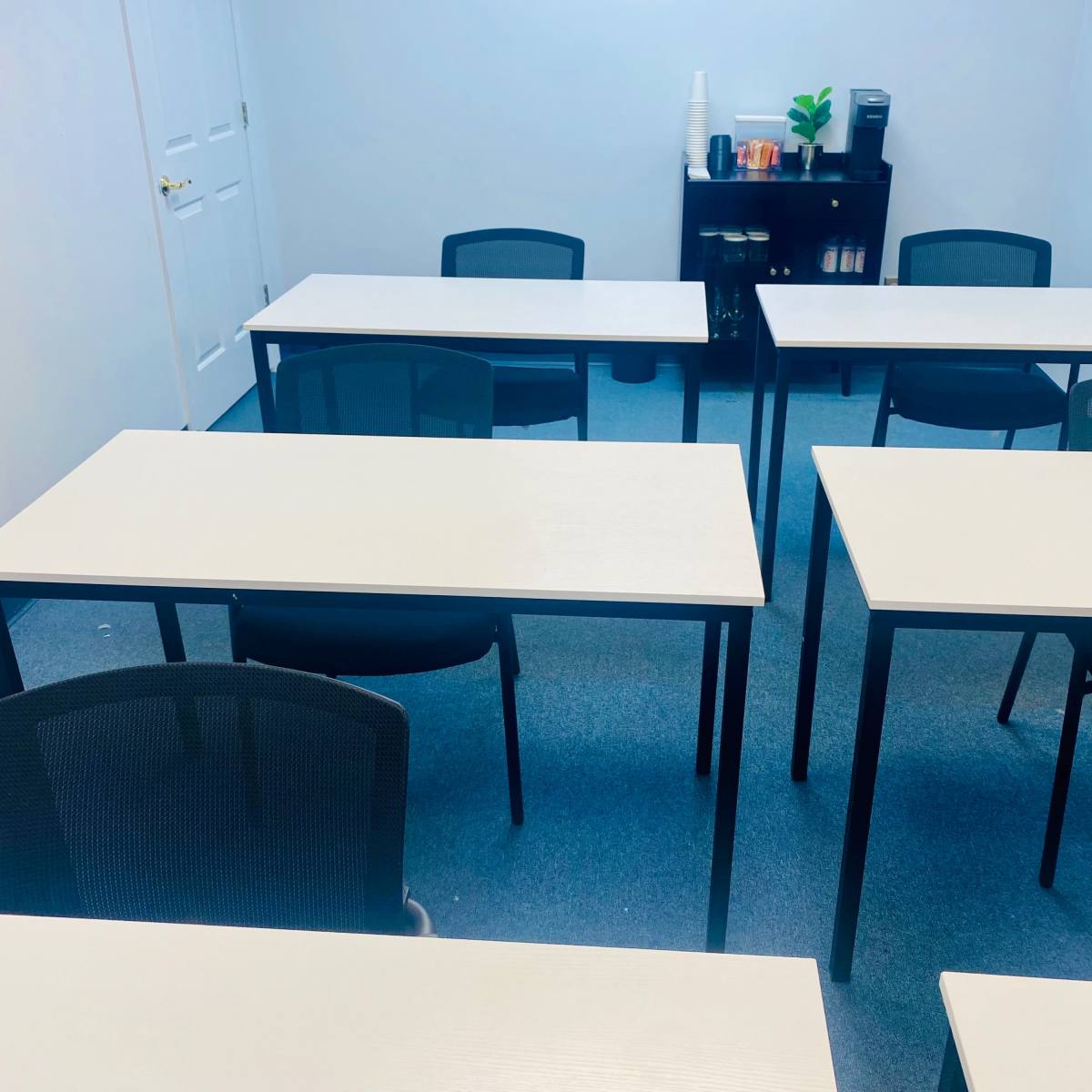 A brightly lit classroom with several empty tables and chairs arranged neatly on a blue carpeted floor, alongside a small shelf holding various items.