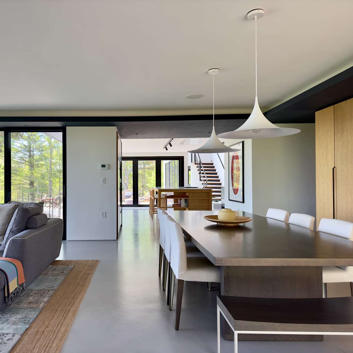 A modern living space featuring a gray sofa, a long dining table, and large windows with a view of greenery outside.
