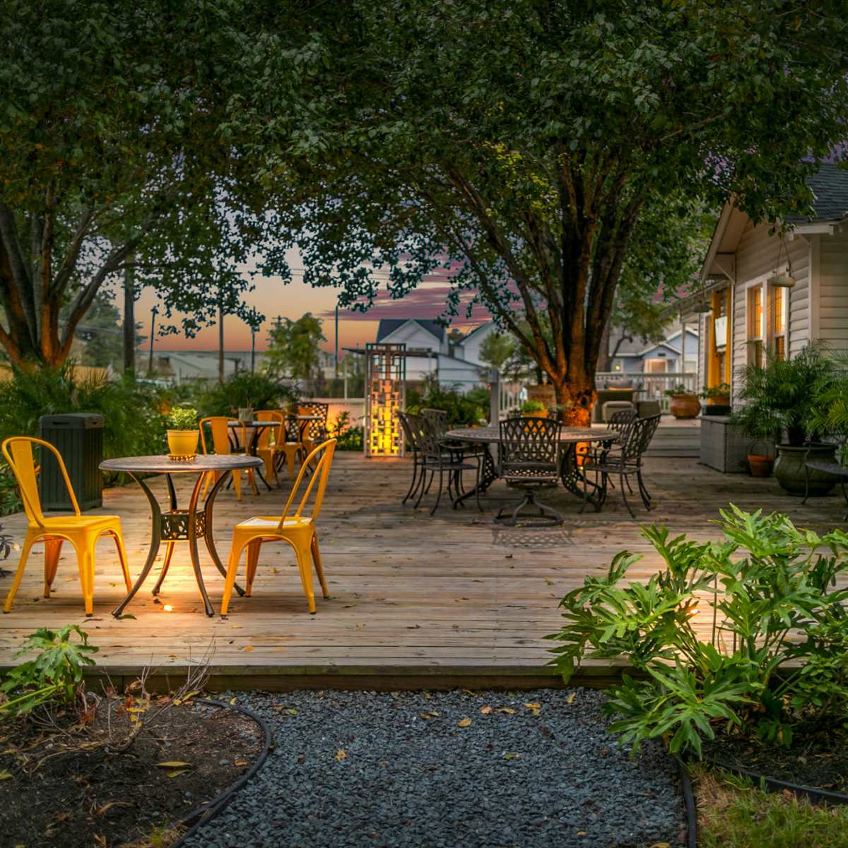 A wooden deck with several tables and chairs surrounded by plants and trees, illuminated by warm lighting from the windows of a nearby house.
