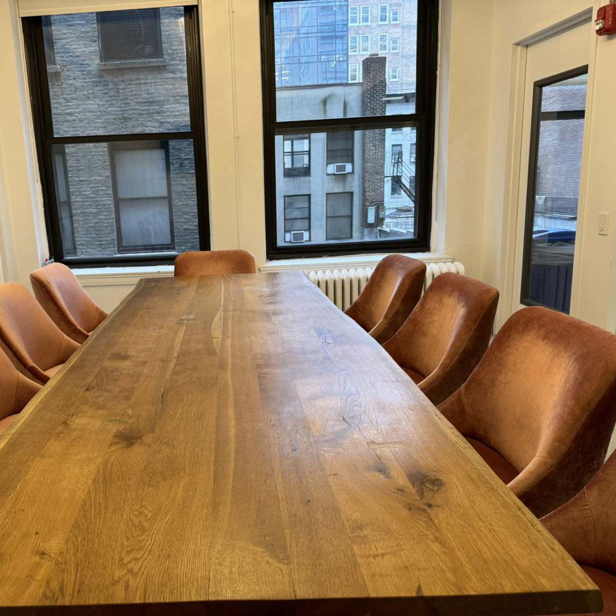 A long wooden conference table surrounded by eight upholstered chairs in a bright, modern room with large windows.