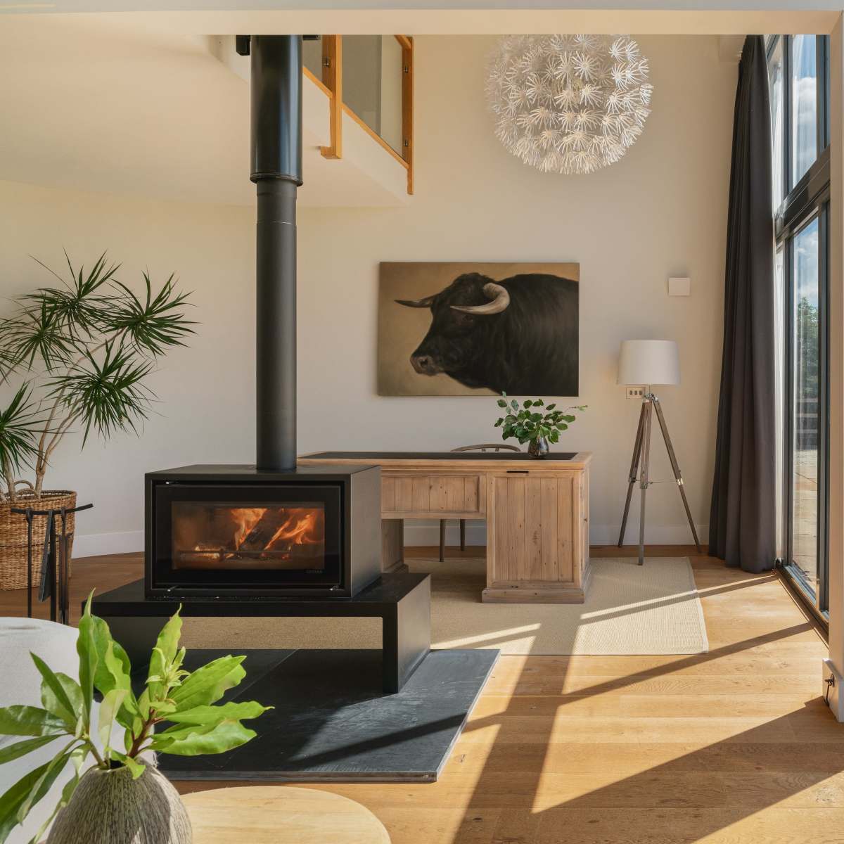 A modern living space features a wood stove, a wooden desk, a large artwork of a bull on the wall, and large glass doors that open to a patio area.