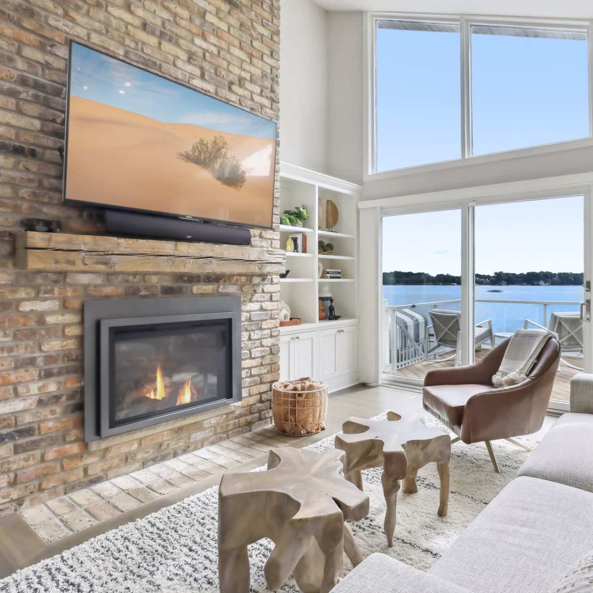 A modern living room with a brick wall, a large TV above a fireplace, and large windows overlooking a lake.
