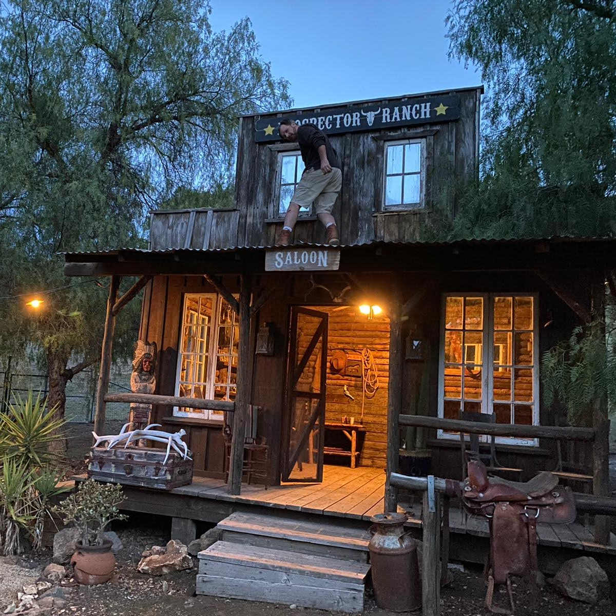 A rustic wooden saloon building with a balcony and a figure sitting on the roof is illuminated by warm lights at dusk.
