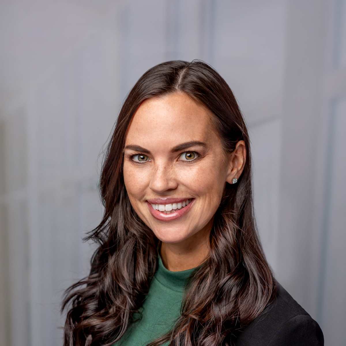 A woman with long, dark hair smiles while wearing a green top and black blazer against a softly blurred background.