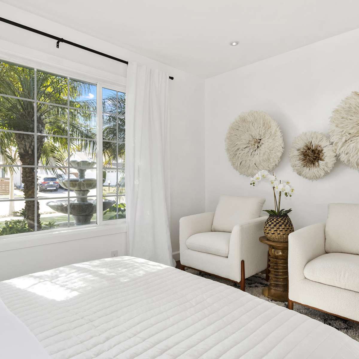 A bright bedroom features two armchairs and decorative wall art, with large windows offering a view of palm trees outside.