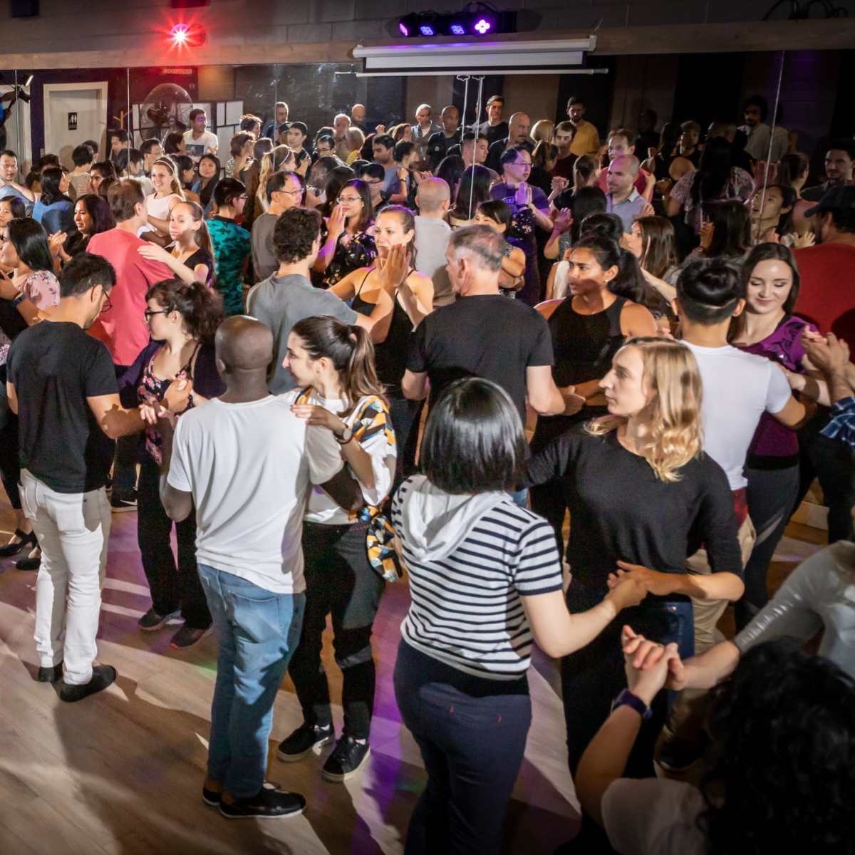 A large group of people dance together in a bustling dance studio with colorful lights and a lively atmosphere.