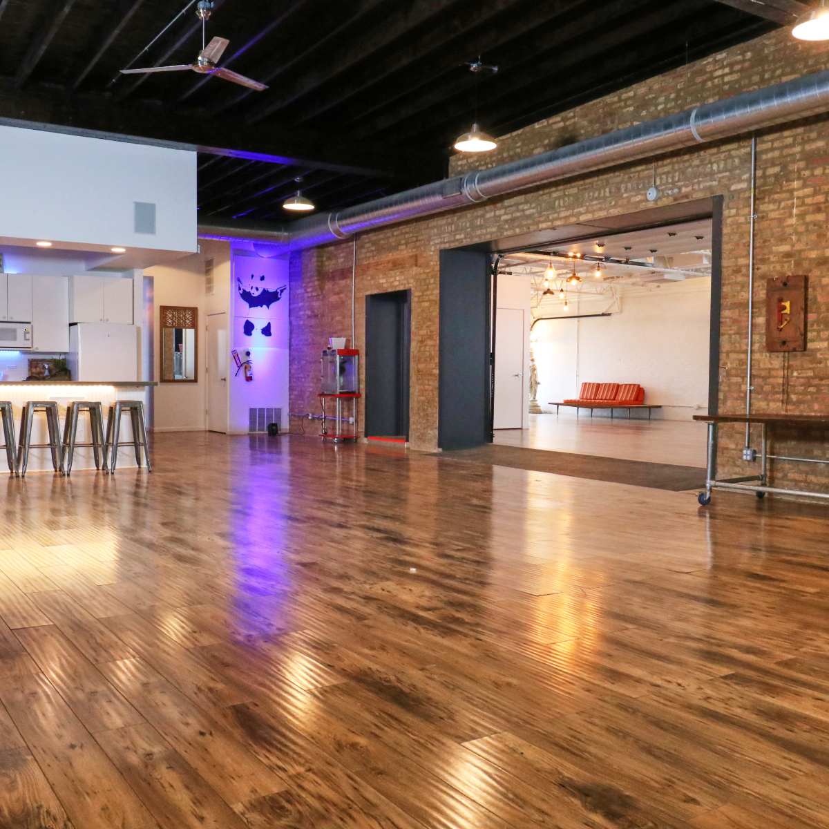 A spacious, industrial-style room features polished wooden floors, exposed brick walls, and a kitchen area with bar stools and modern fixtures.