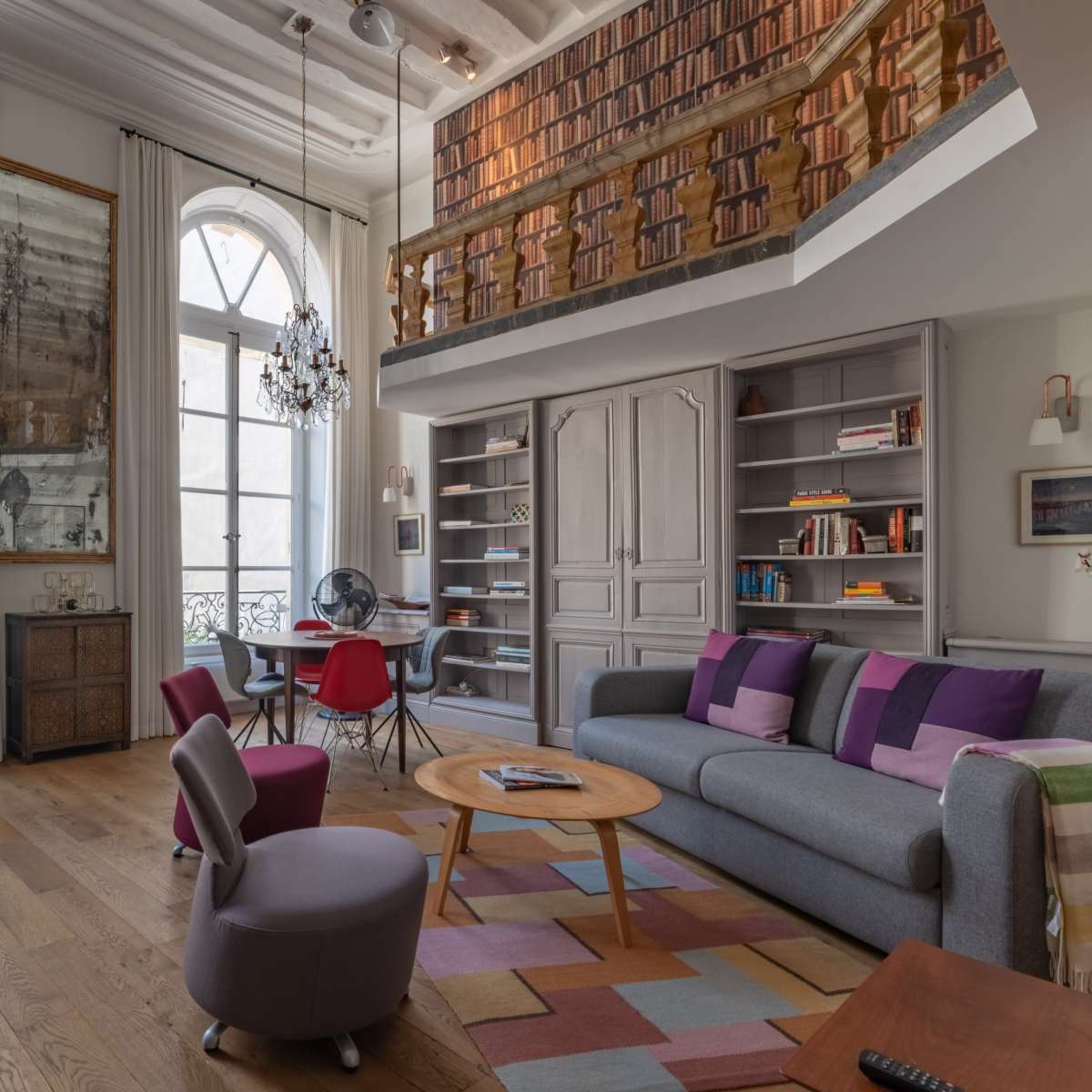 A modern living room with two large windows, a cozy seating area, and a lofted bookshelf overhead.