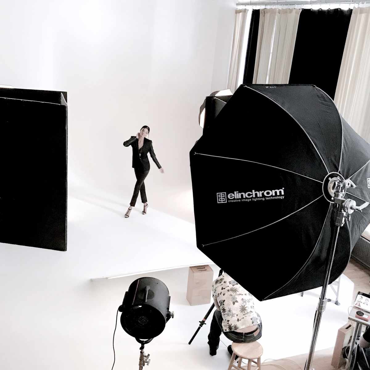 A model poses on a white backdrop during a photoshoot, while a large softbox and studio lights illuminate the scene.