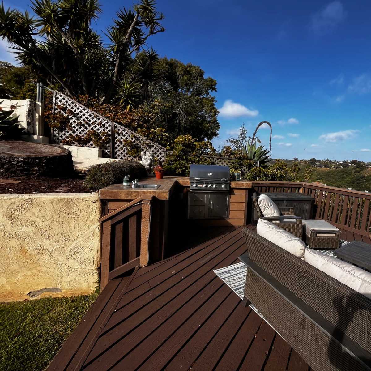 A wooden deck features a grill and outdoor seating, overlooking a green hillside under a blue sky.