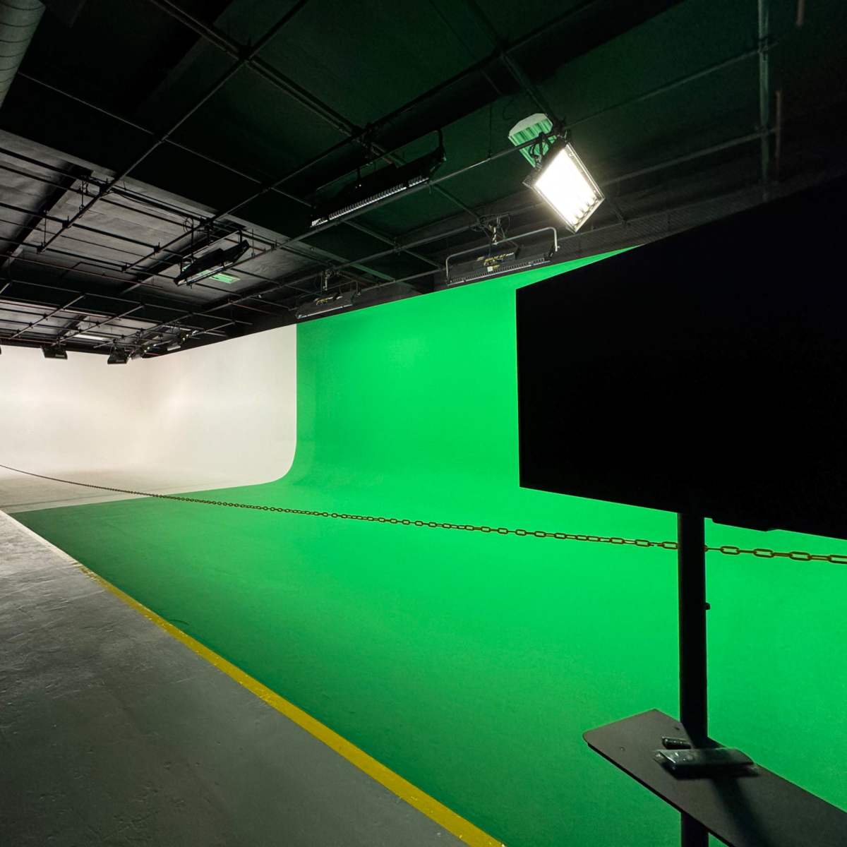 A spacious studio with a green and white backdrop, a low table with a monitor, and chairs visible in the background.