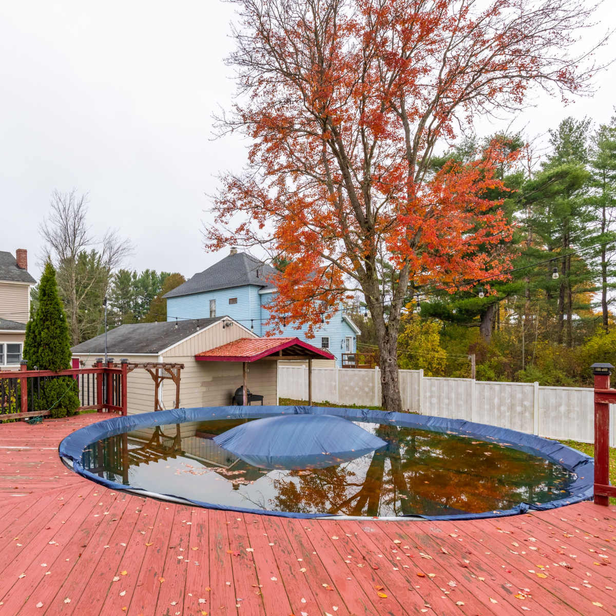 The scene shows a backyard deck with a covered pool and nearby houses, surrounded by trees displaying autumn foliage.