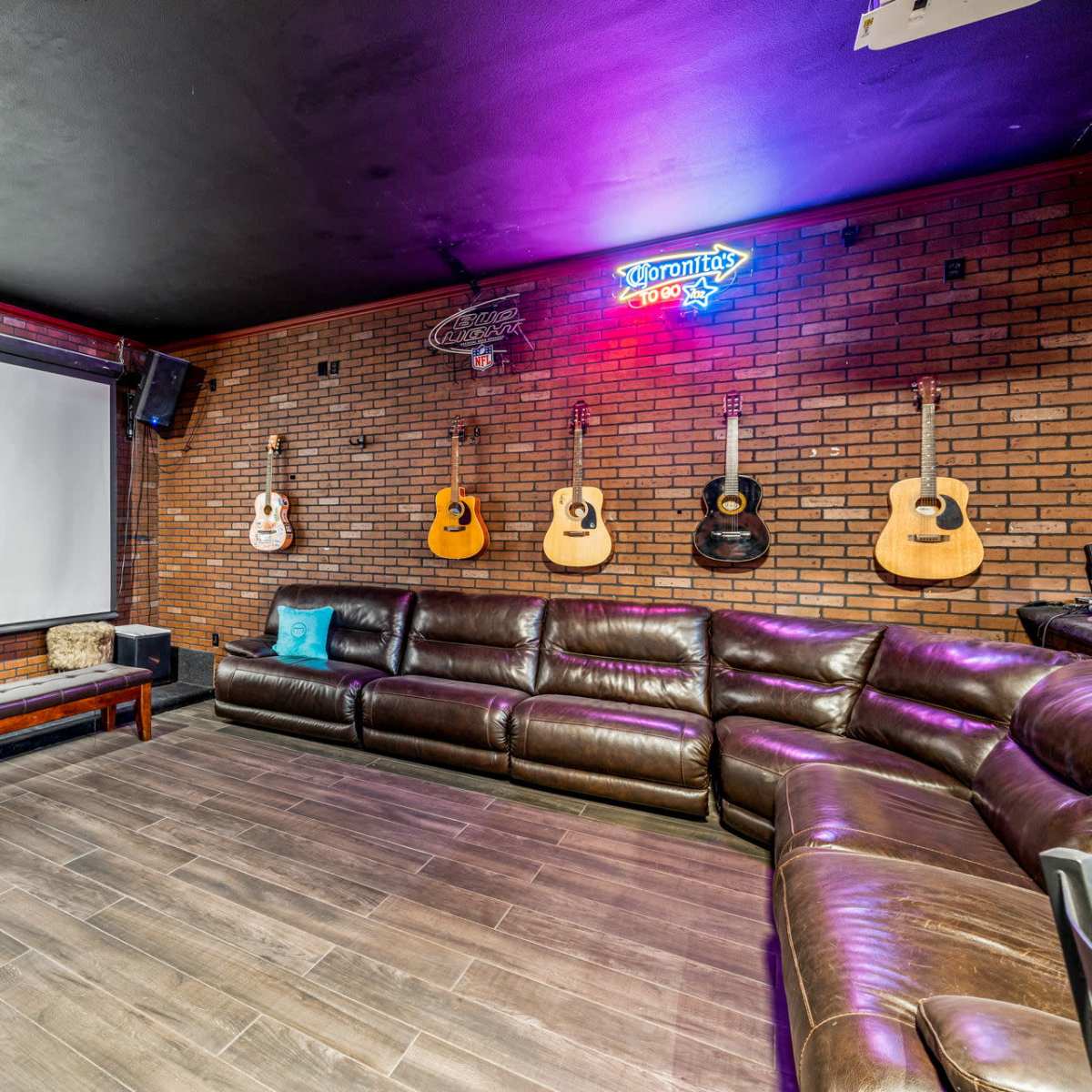A lounge area with a large screen at one end, surrounded by a brick wall and several acoustic guitars hanging above a leather sectional sofa.