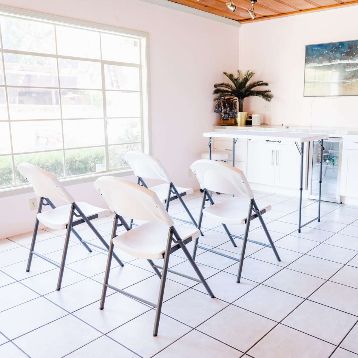 A bright room features a row of six folding chairs facing a small counter with a modern painting on the wall.
