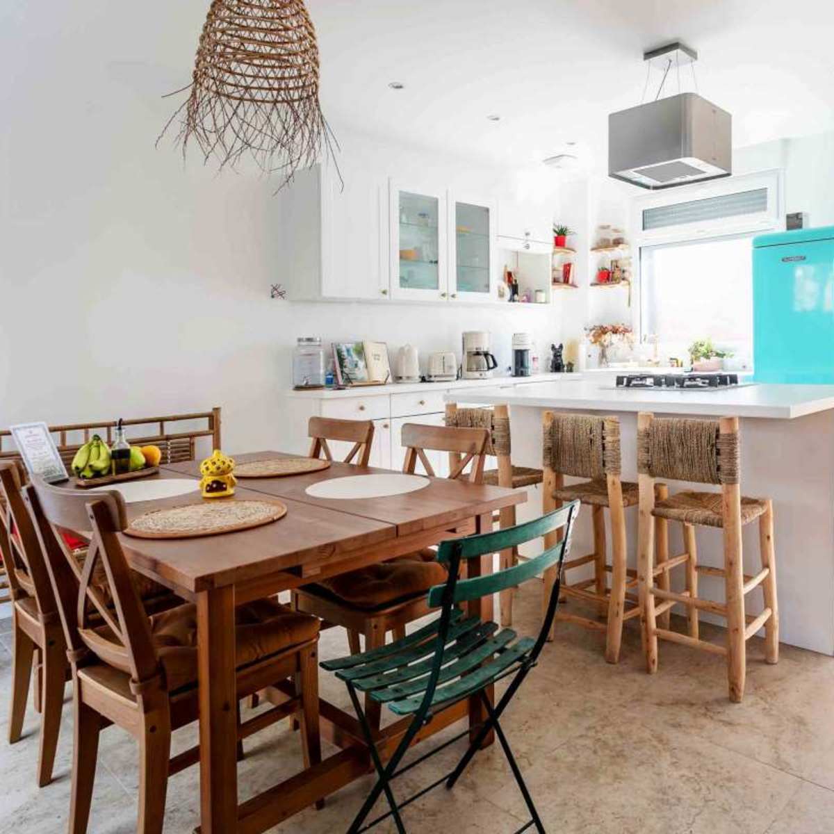 A modern kitchen features a wooden dining table surrounded by chairs, a light blue refrigerator, and an adjacent outdoor area visible through a sliding door.