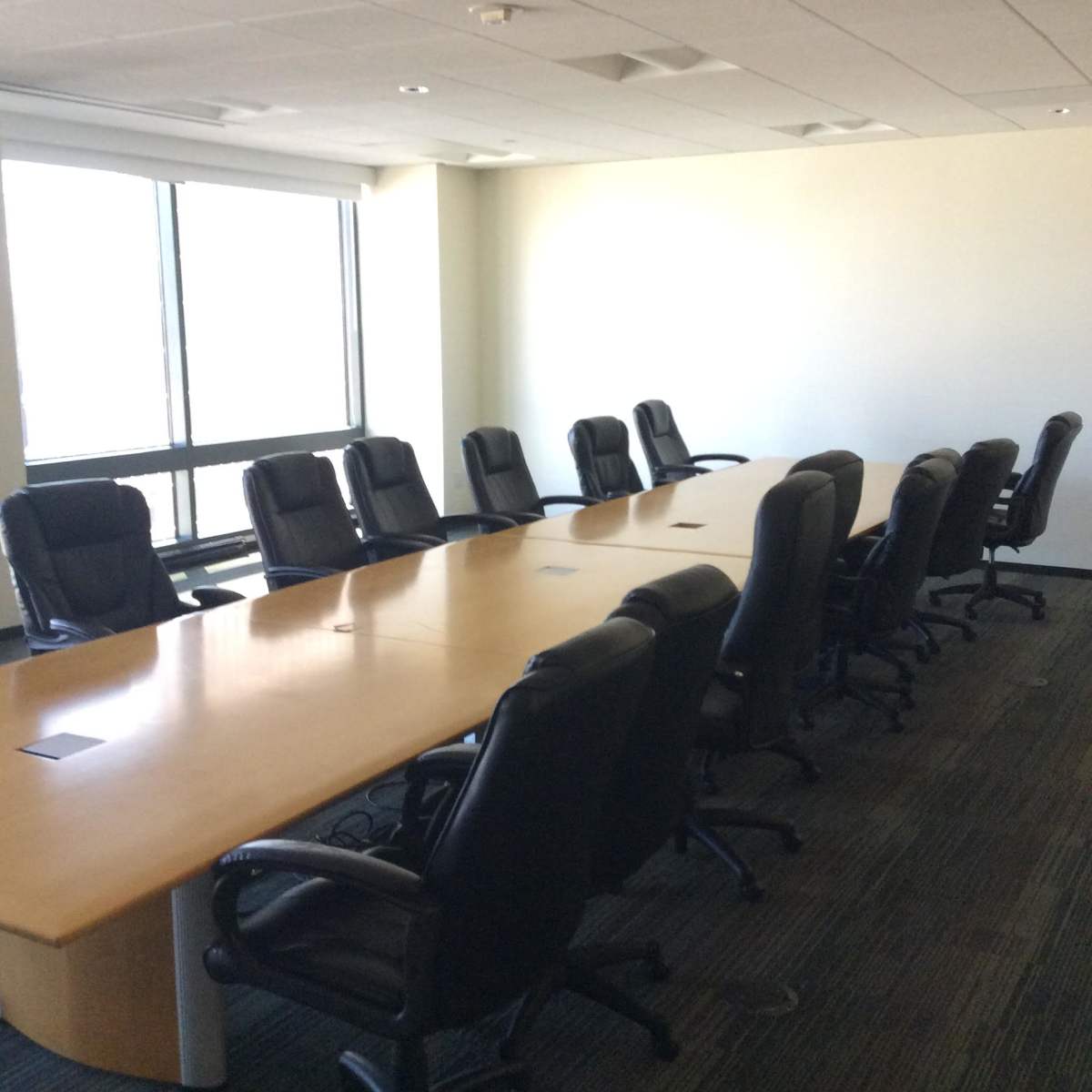 A large conference room features a long wooden table surrounded by black ergonomic chairs and a large window providing natural light.