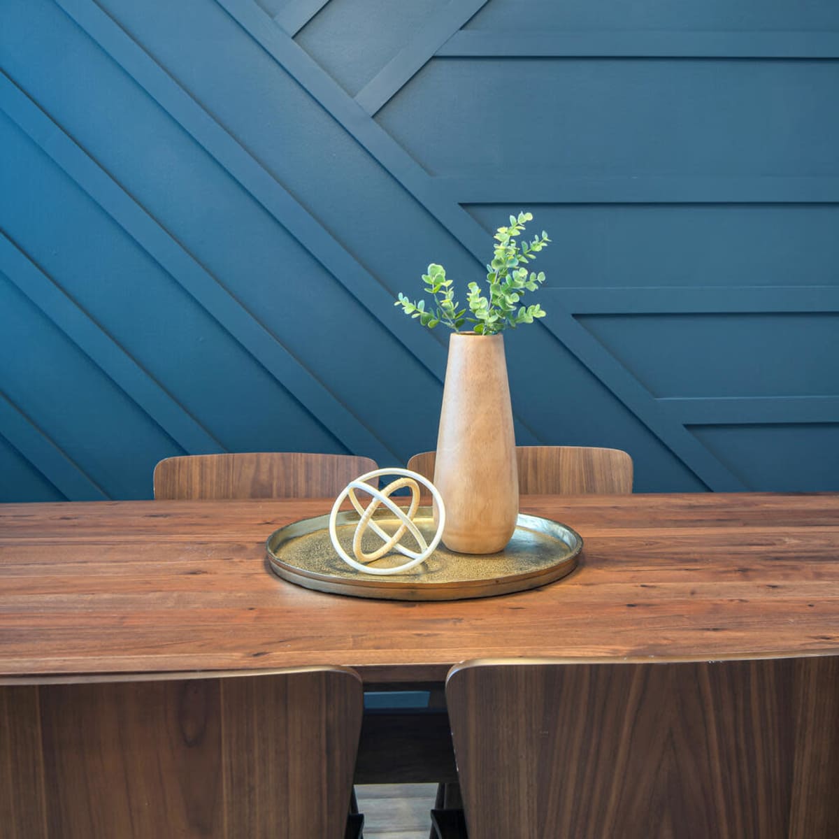 A wooden table is set with a round tray holding a decorative item and a vase containing green foliage, against a blue geometric-patterned wall illuminated by pendant lights.
