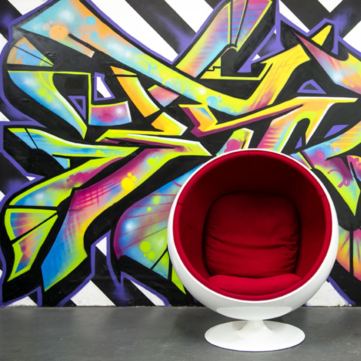 A round red chair is positioned in front of a vibrant, multicolored graffiti wall featuring angular designs.