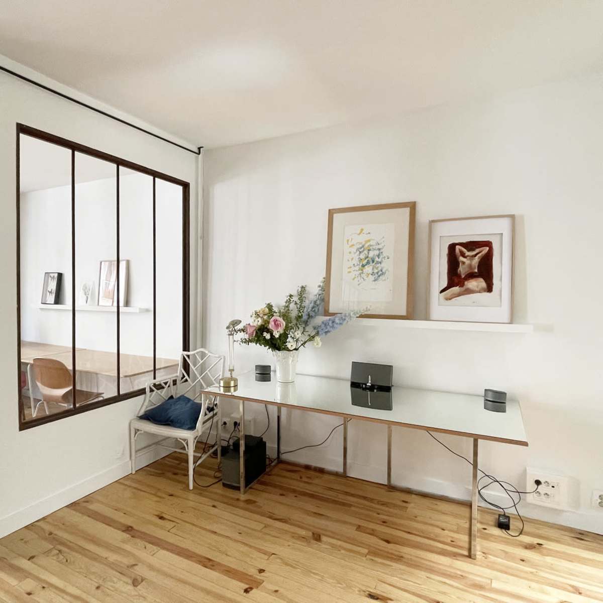 A modern workspace with a glass desk, decorative artwork on the wall, and a floral arrangement placed on the desk, adjacent to a partition with windows.