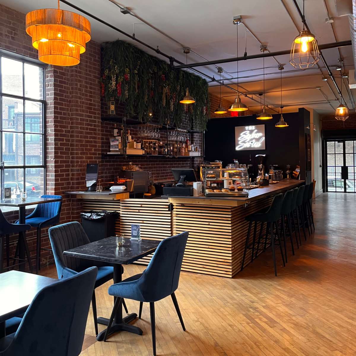 A modern café interior with wooden floors, a bar area adorned with hanging lights, and a mix of seating options including dark chairs and a black table.