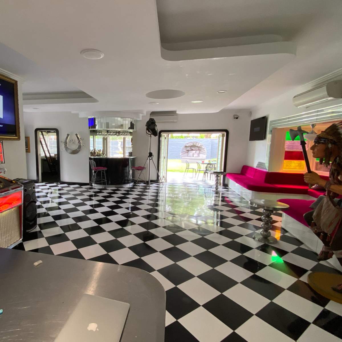 A modern room with checkered black and white flooring, a jukebox, and a red seating area, featuring large windows that open to a garden view.