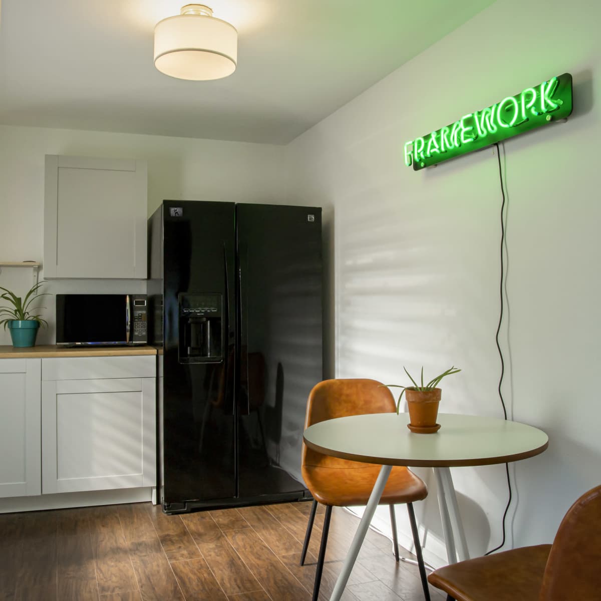 A modern kitchen features a black refrigerator, a microwave, a small table with two chairs, and a neon sign on the wall.