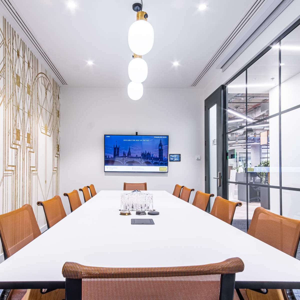 A conference room features a long table surrounded by chairs, with a screen mounted on the wall and large windows displaying a yellow accent wall.