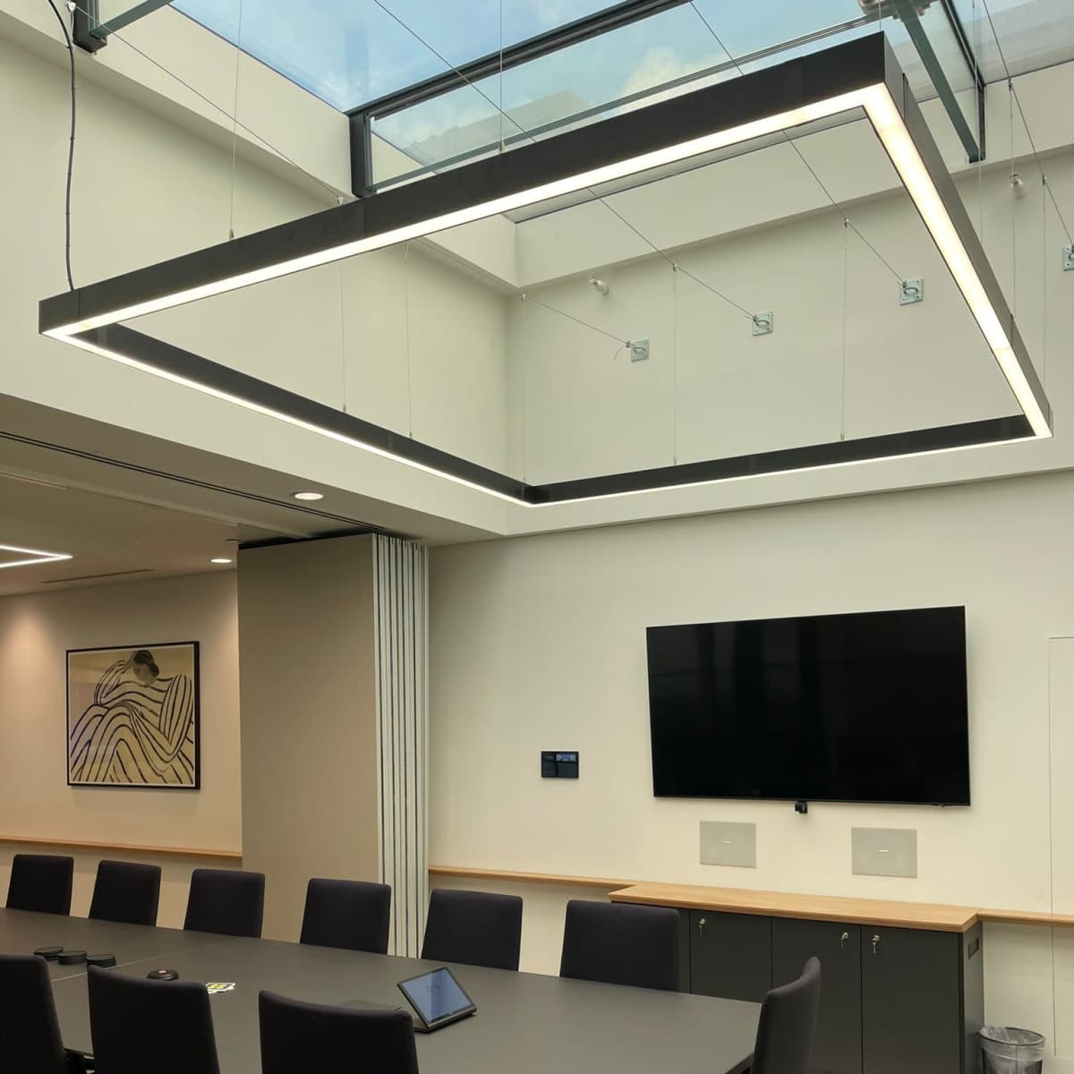 A modern conference room features a large rectangular table surrounded by black chairs, with a ceiling-mounted light fixture and a screen on the wall.