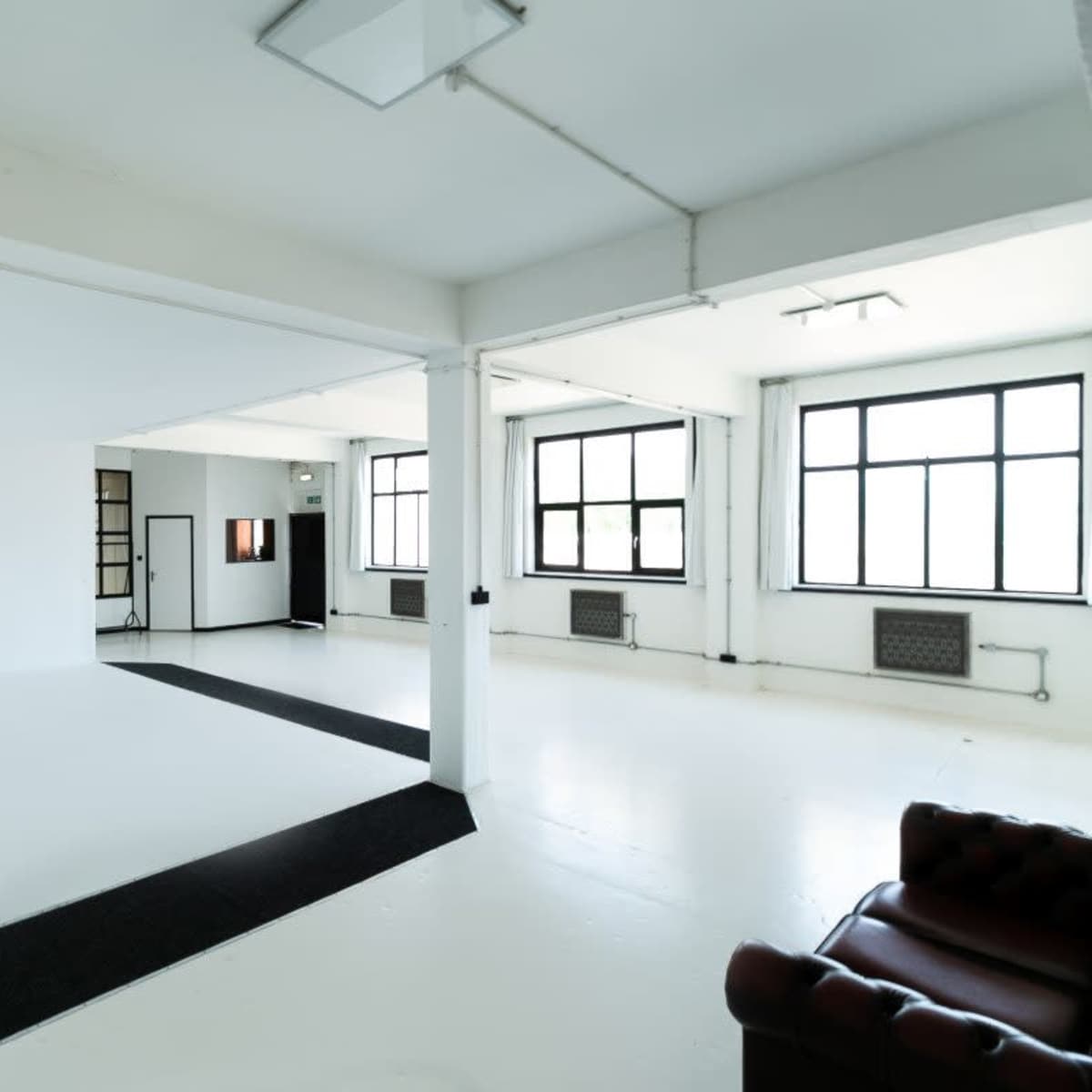 A spacious studio with white walls, large windows, and a black carpet along the floor, featuring a brown leather couch in one corner.