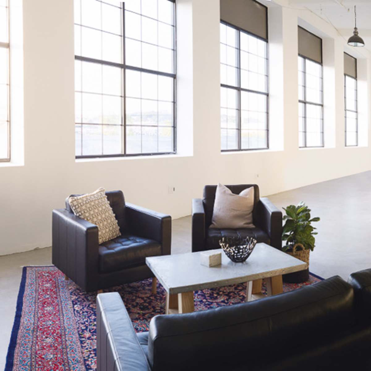A spacious, modern interior with a seating area featuring black couches, a coffee table, and a dining table set for six, illuminated by large windows.