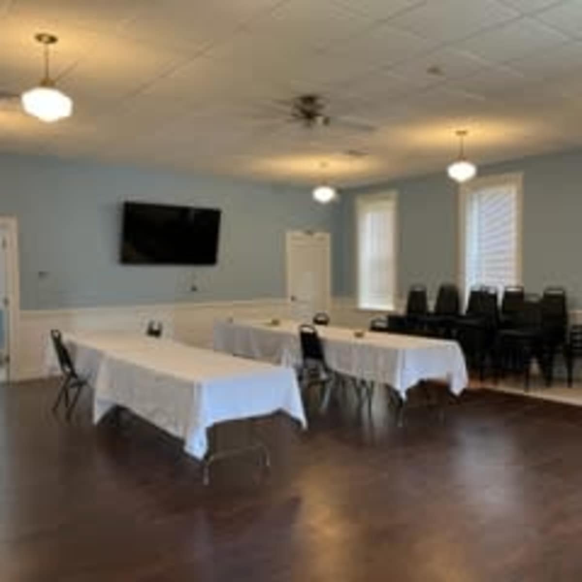 A well-lit room features several white-covered tables arranged in the center, with black chairs stacked against the wall and a television mounted on the front wall.