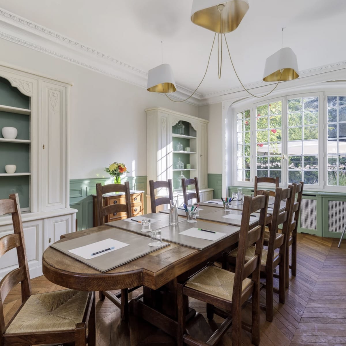 A well-furnished meeting room features a wooden table surrounded by chairs, with a large window allowing natural light and shelves displaying decorative items.