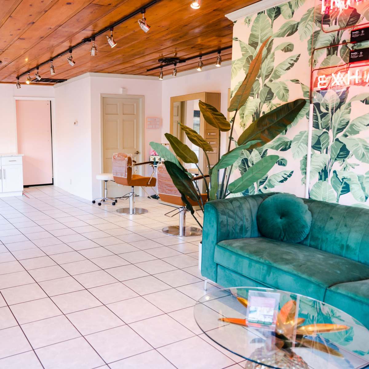 A salon interior with tiled flooring, a green sofa, a coffee table, and a kitchenette area against a backdrop of tropical-themed wallpaper.