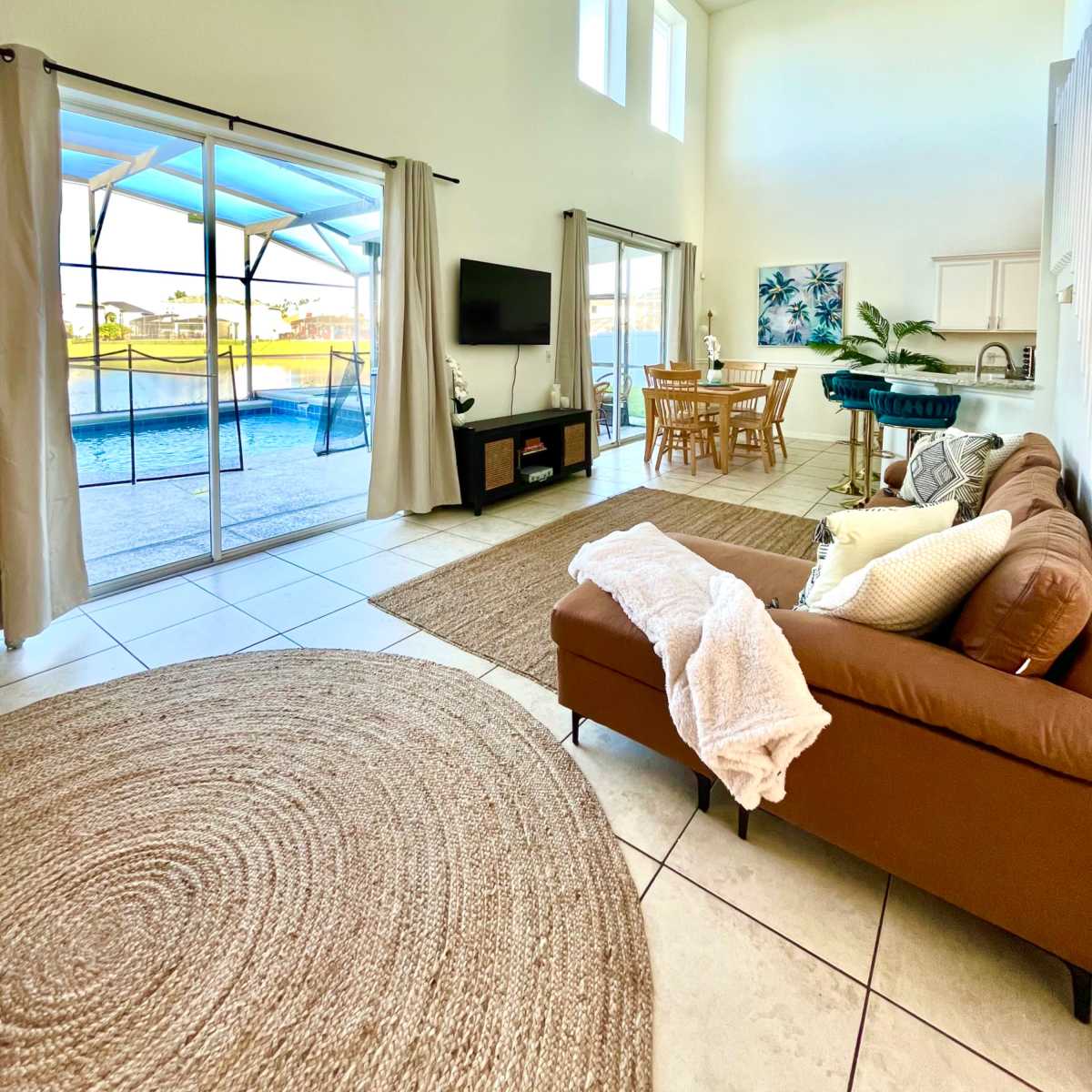 A spacious living room with a brown sofa, a round rug, a dining area, and large windows offering a view of an outdoor pool.
