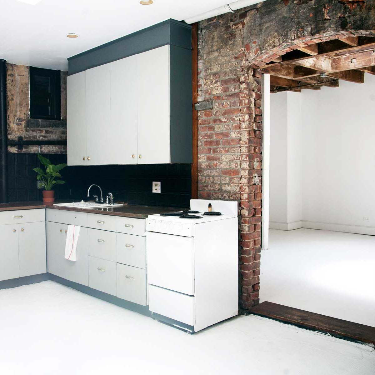 A kitchen with minimalist cabinetry, a white stove, and exposed brick walls, leading to an empty, light-filled room.