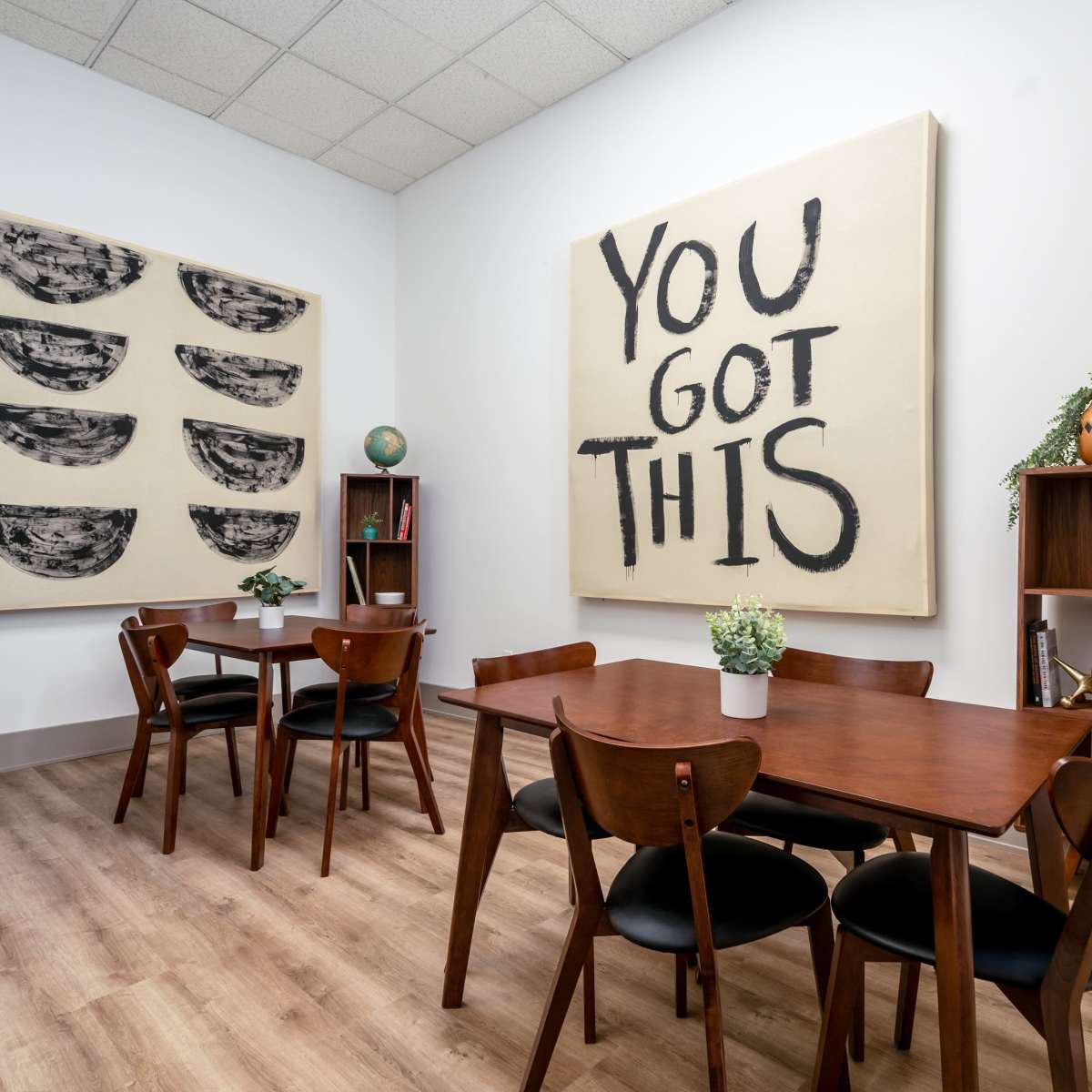 A brightly lit room features wooden tables and chairs arranged around the space, with large artworks on the walls and decorative plants on the shelves.