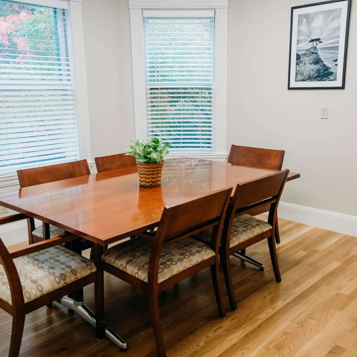 A wooden dining table with six chairs is positioned in a well-lit room featuring bay windows and a framed picture on the wall.