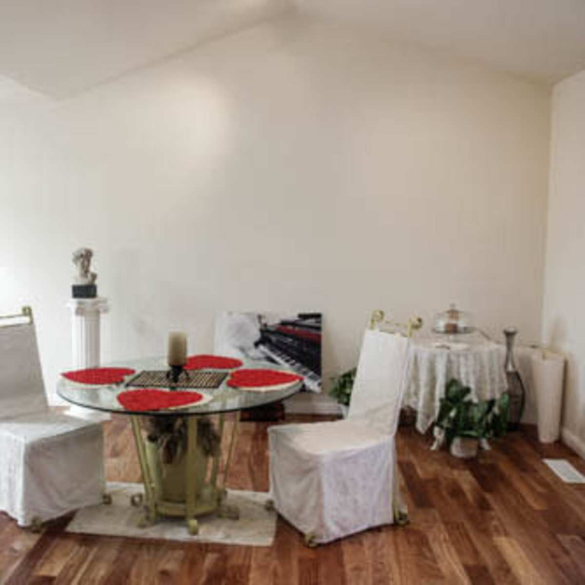 A round table with red placemats is set in a sparsely decorated dining area featuring white chairs and wooden flooring.