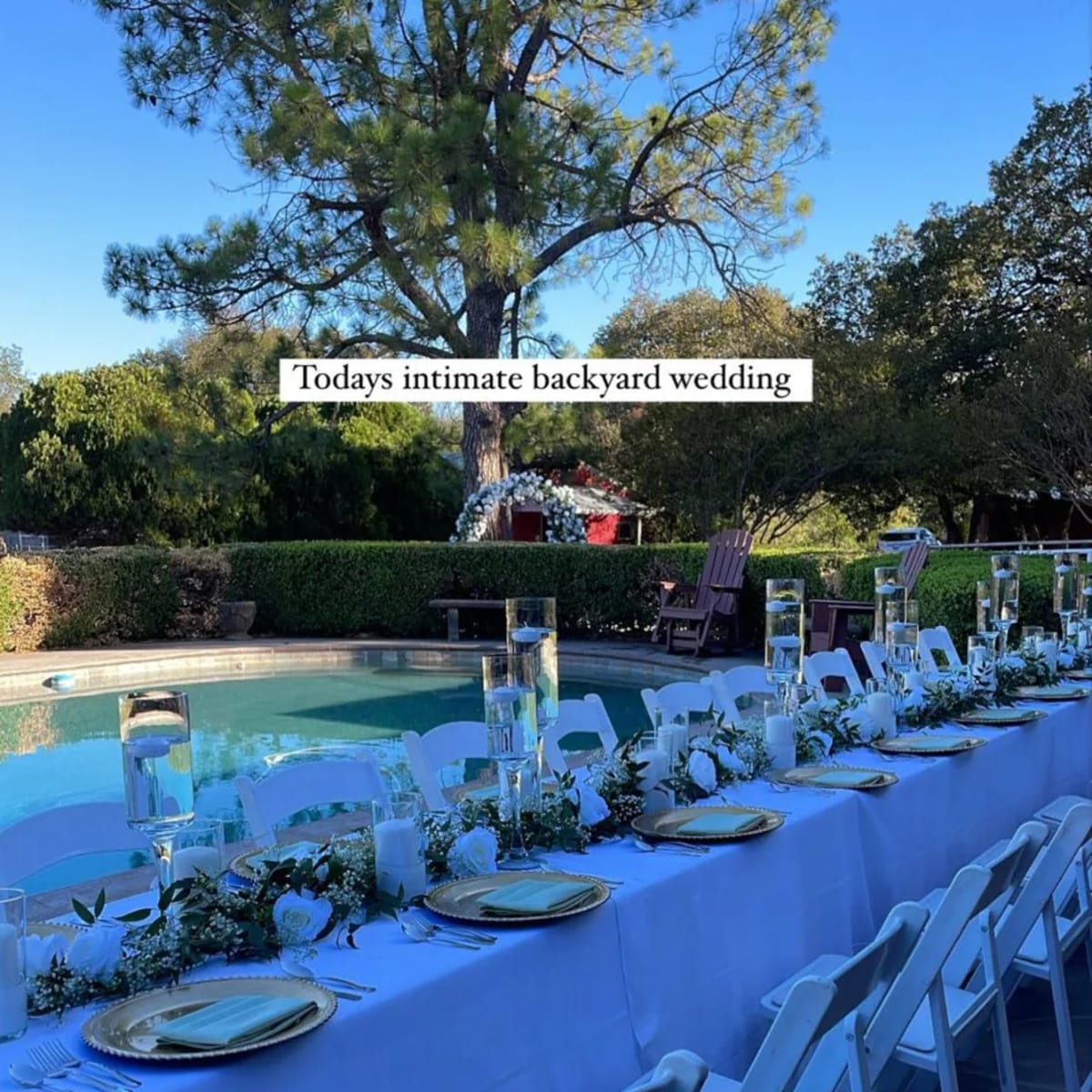 A long table set for an outdoor wedding by a pool, adorned with floral decorations and glass candle holders.