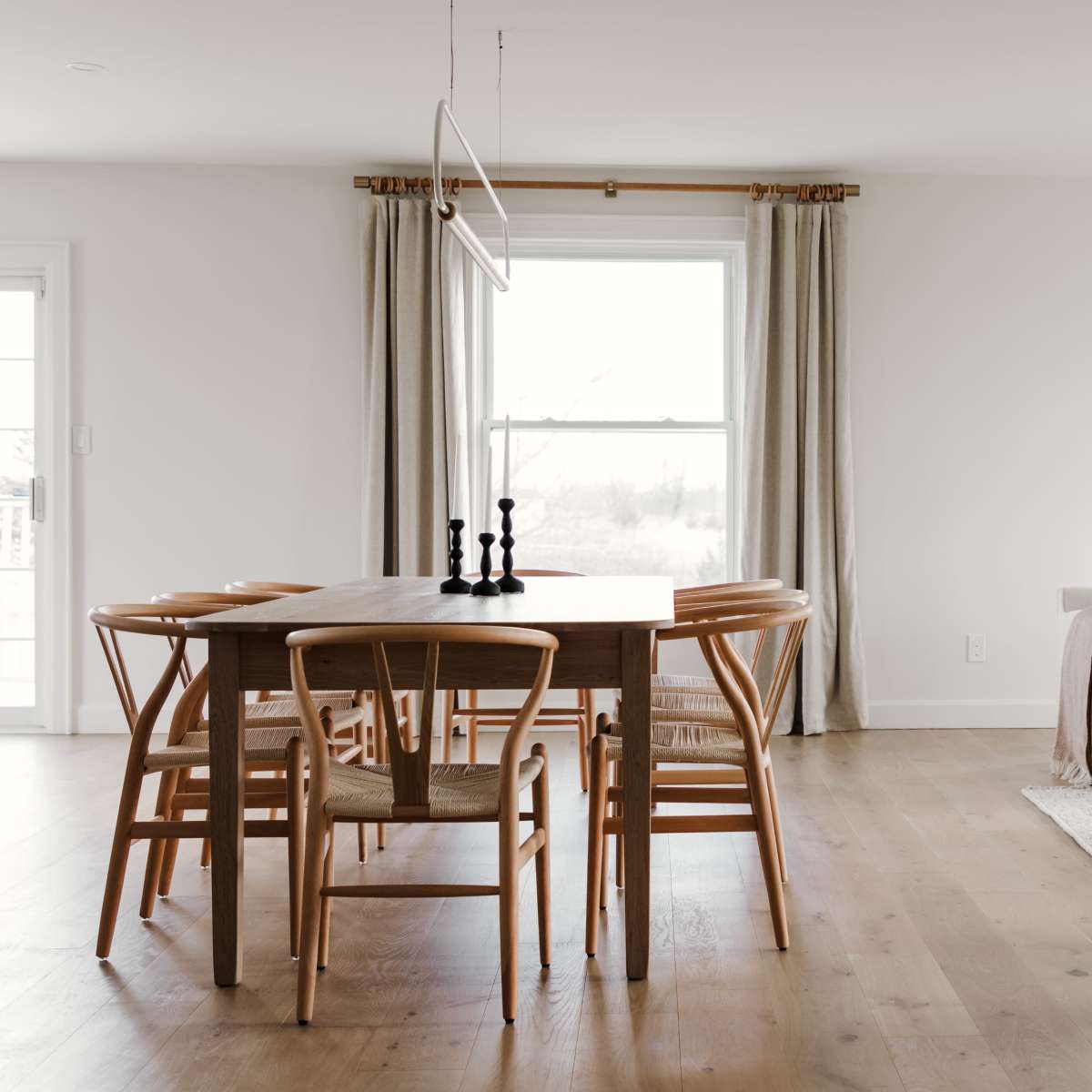 A modern dining area with a wooden table surrounded by six chairs, a sofa, and large windows draped with light curtains.