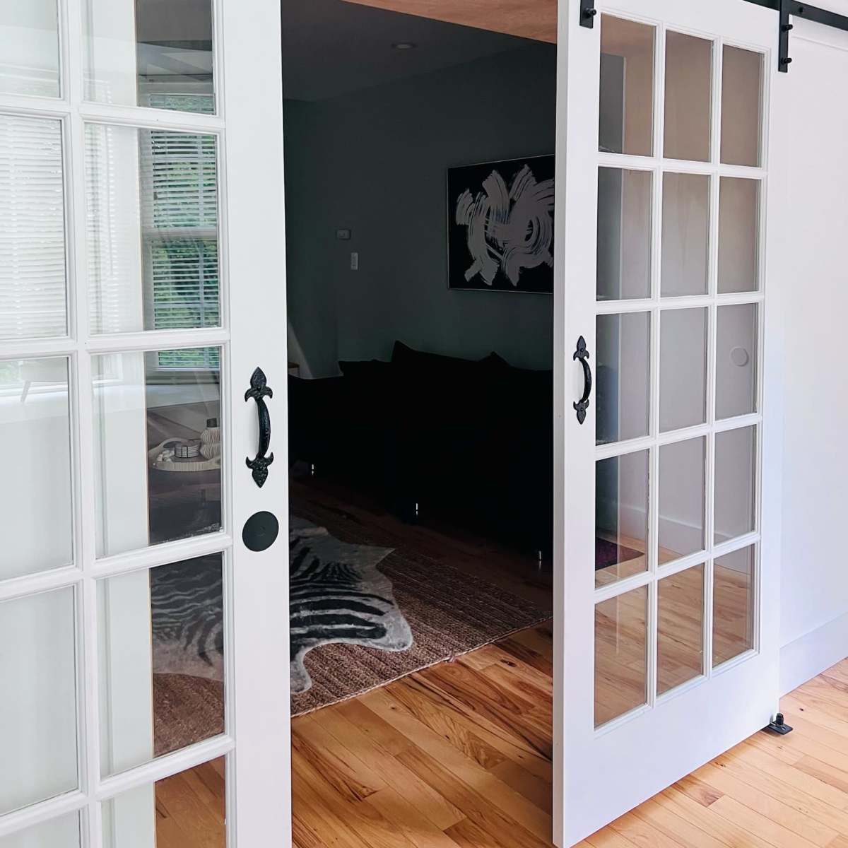 A set of white double barn doors with glass panels, mounted on a black sliding track, leading to a furnished room with a dark couch and a patterned rug.