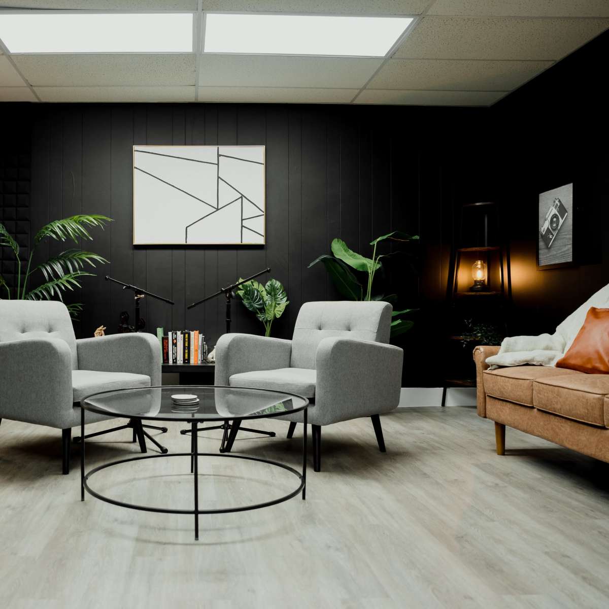 A modern, minimalist room featuring two gray armchairs facing a circular glass coffee table, with plants and a bookshelf in the background.