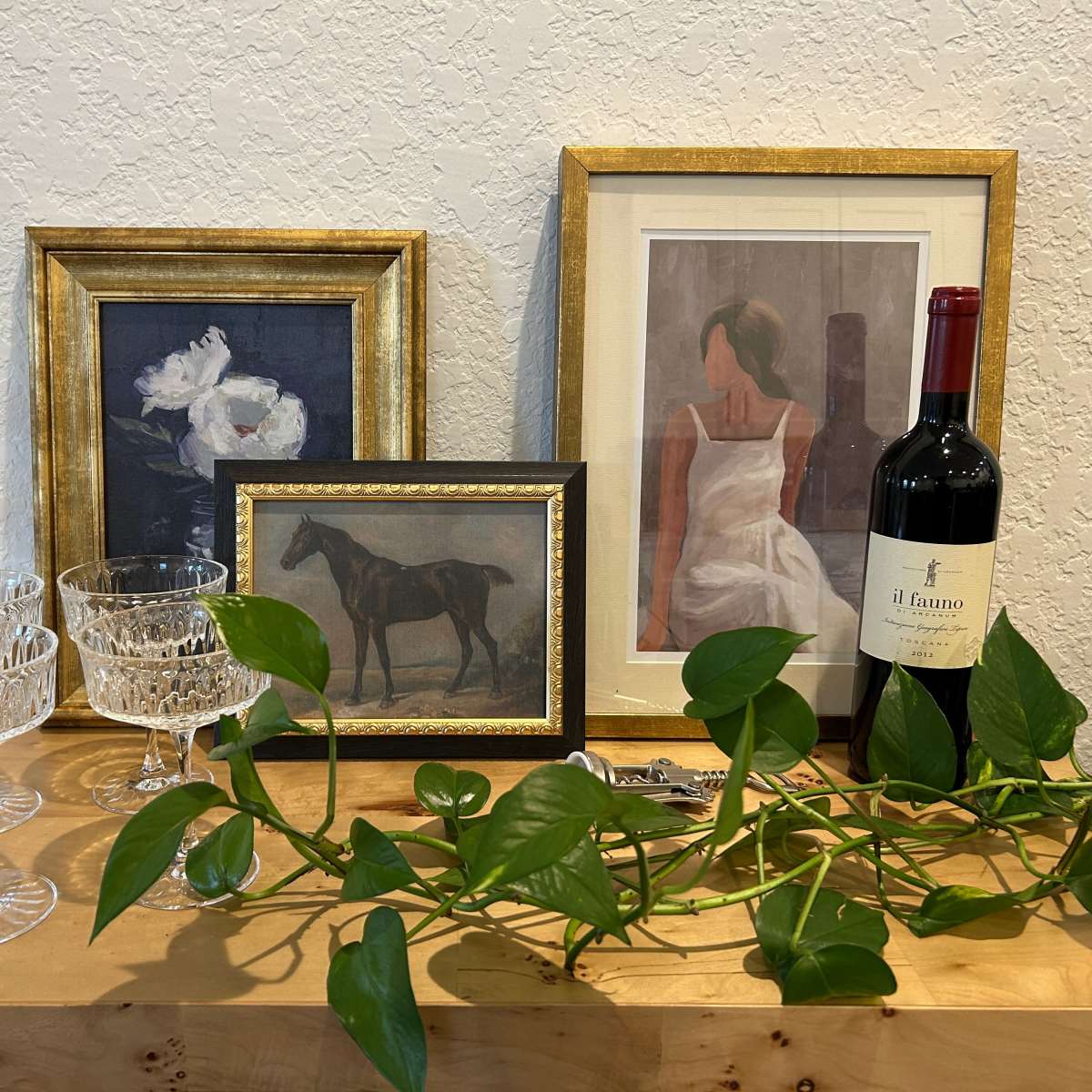 A wooden table displaying three framed artworks, two crystal glassware pieces, a bottle of wine, and a green vine.