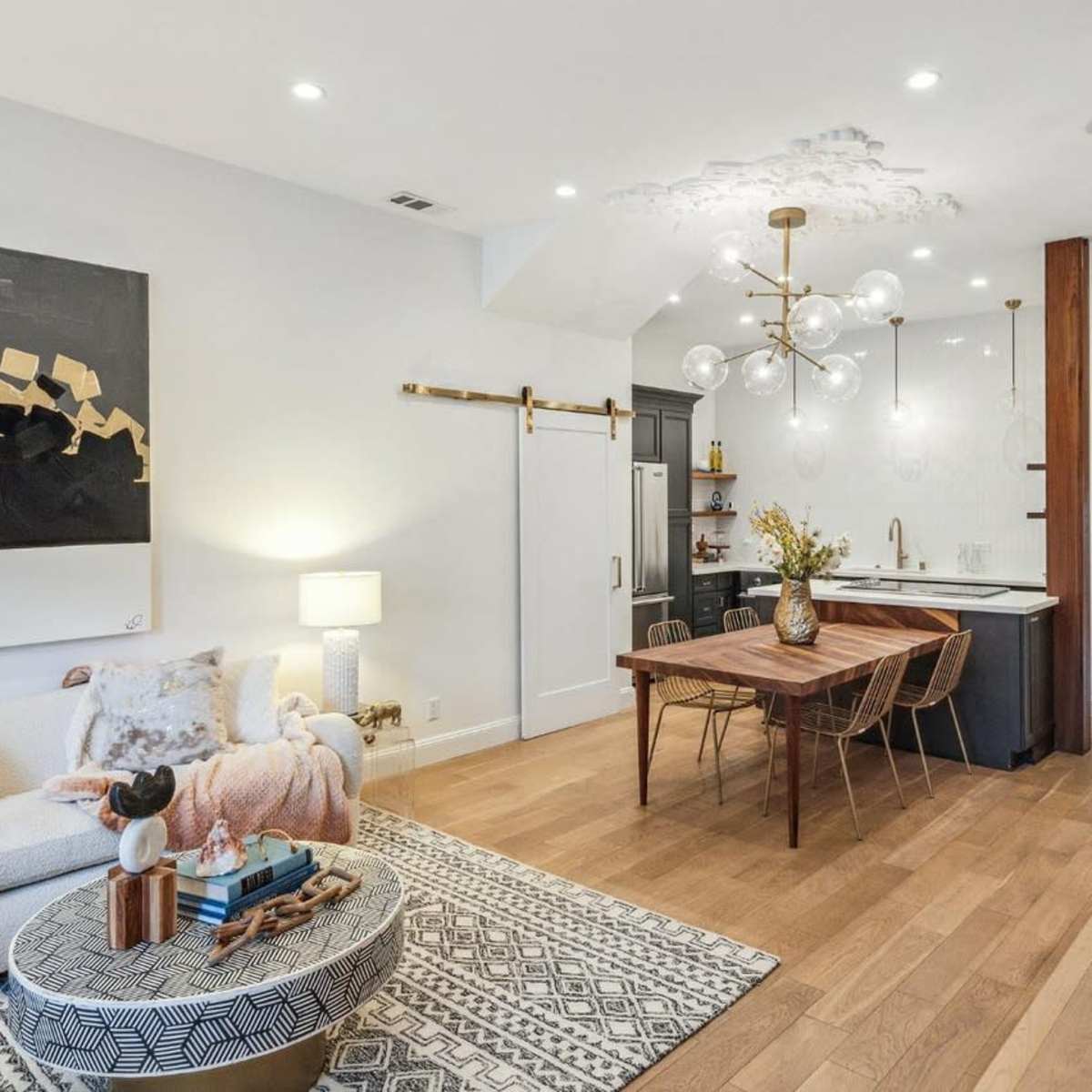 A modern living space featuring a white sofa, a round coffee table with decor, and a dining area with a wooden table and stylish light fixture, adjacent to a kitchen with dark cabinetry.