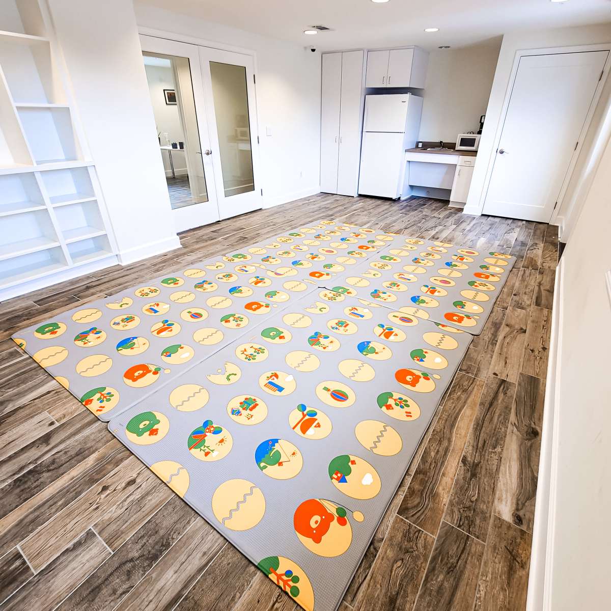 A brightly patterned play mat covering the floor of a kitchen area with white cabinets and appliances in a light-filled room.