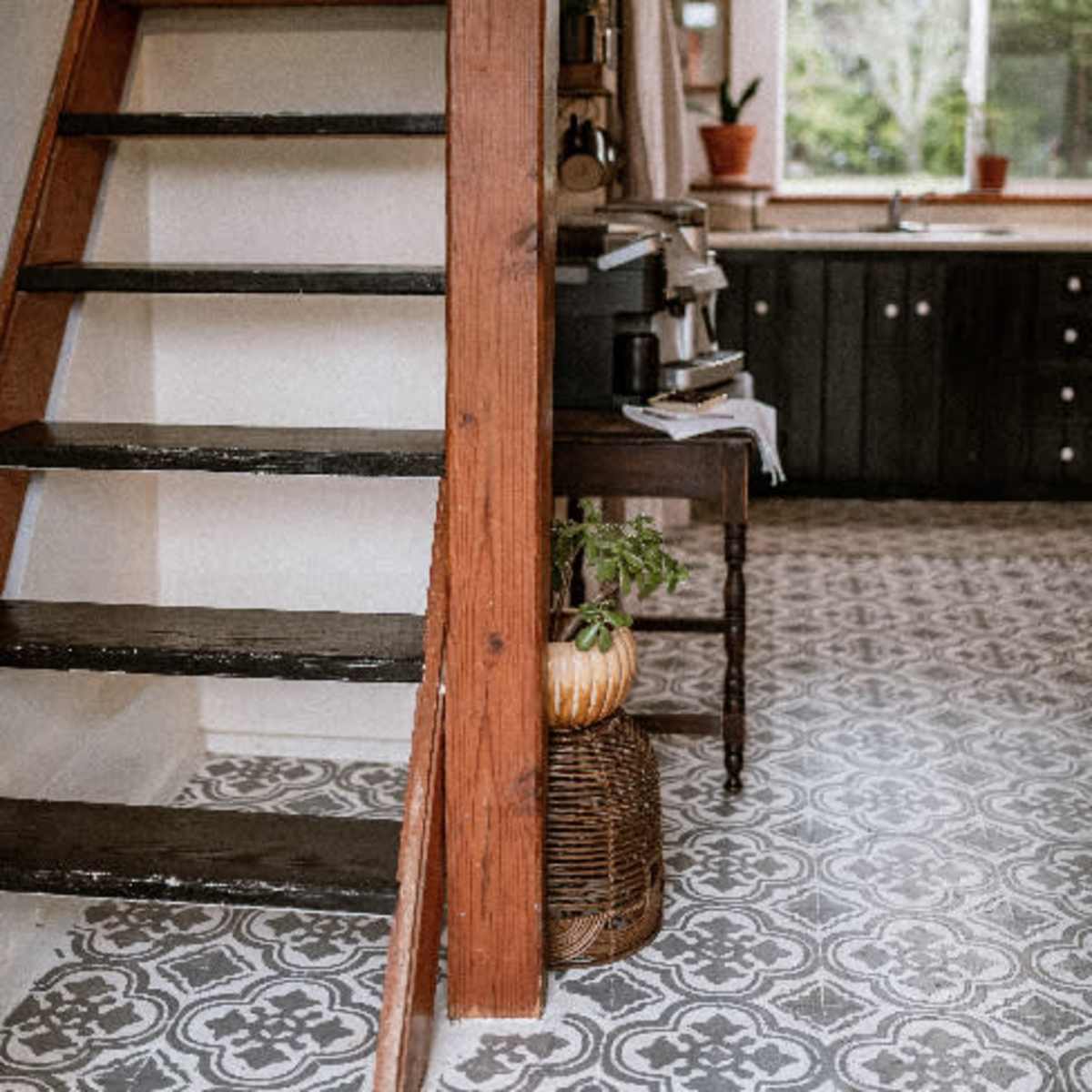 A kitchen area with a staircase leading to an upper level, featuring black cabinetry, a patterned floor, and potted plants.