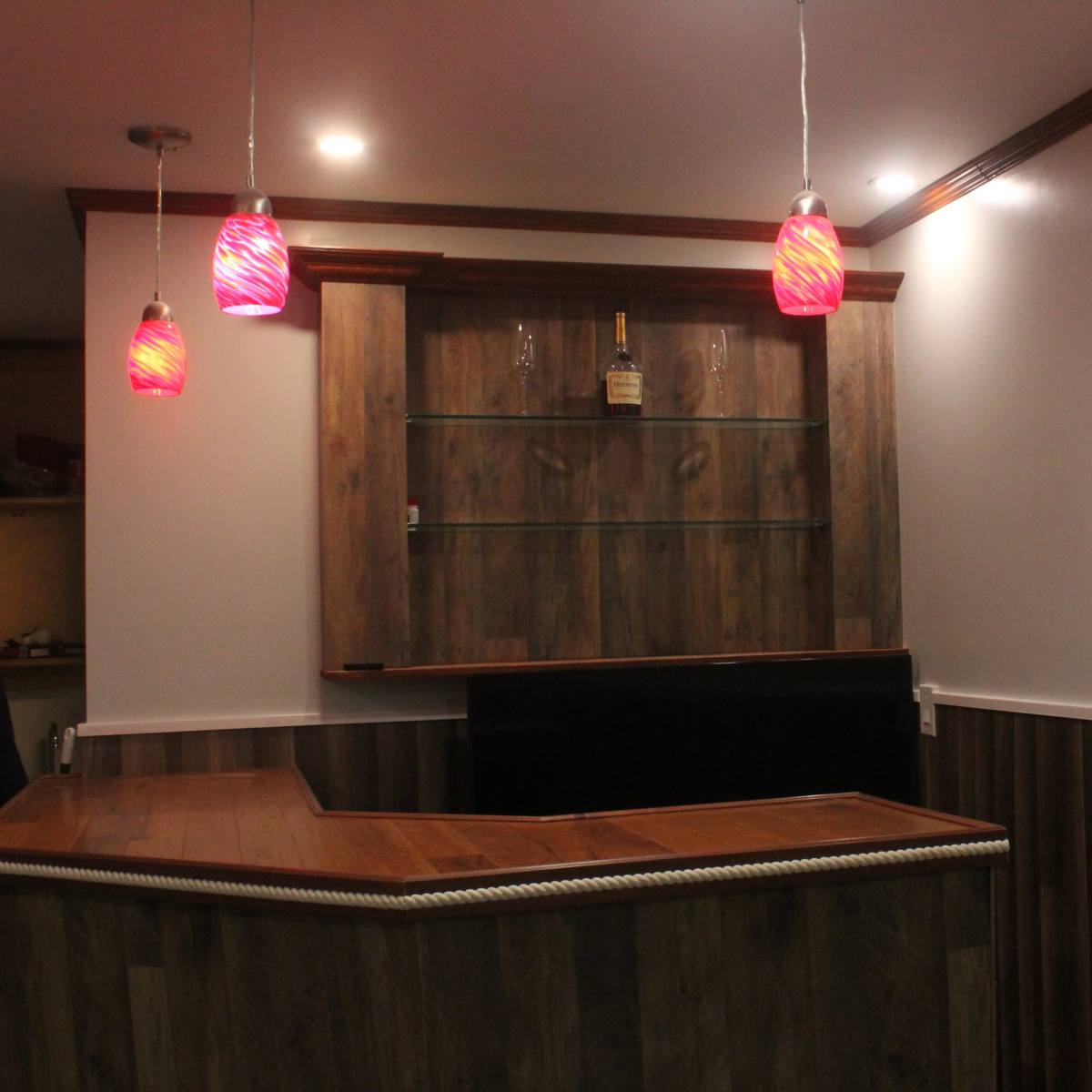 A bar area featuring a wooden countertop, glass shelves, and pendant lights with pink shades.