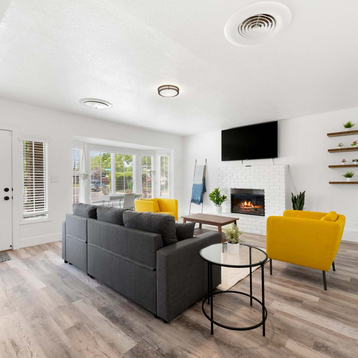 The living room features a gray sofa, yellow chairs, a circular table, a wall-mounted TV, and a fireplace with white brick, all set against a backdrop of large windows.