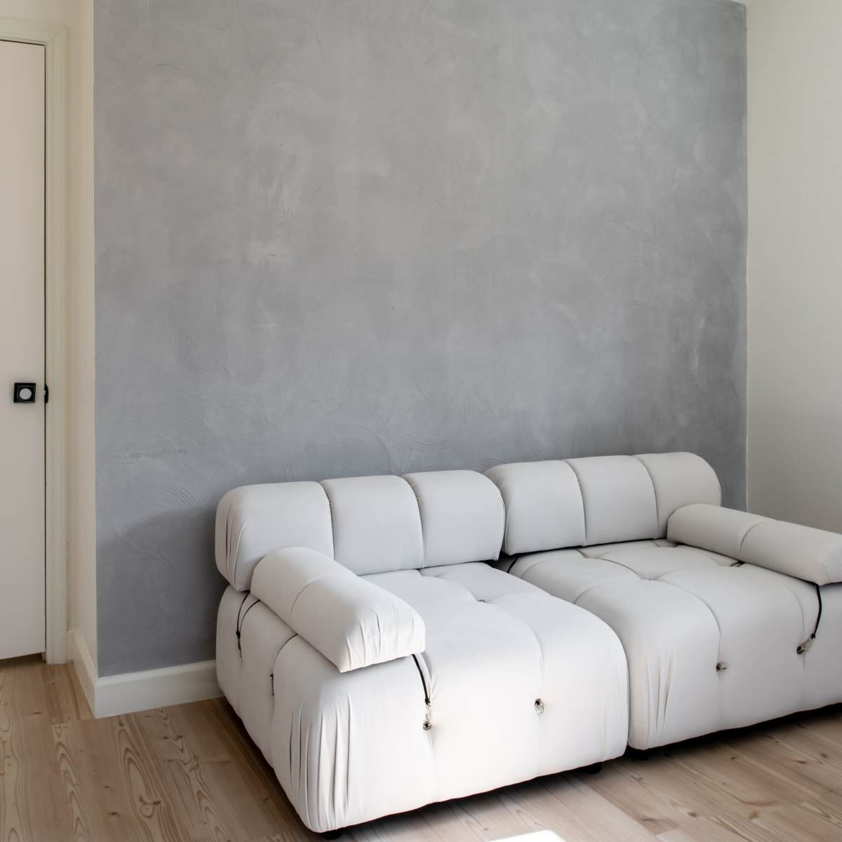 A white, tufted sofa is positioned against a gray wall in a minimalistic room with light wood flooring.